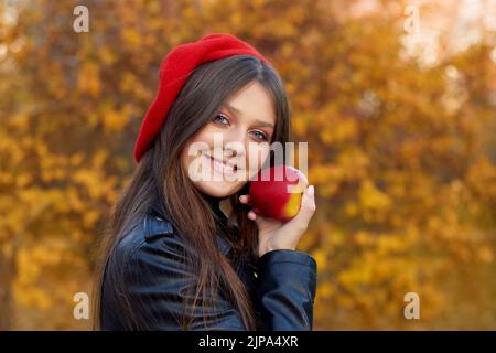 Glückliche, schöne Frau, die rote Baskenmütze trägt, lächelnd direkt auf die Kamera schaut, Apfel isst und Zeit im Freien verbringt Stockfoto