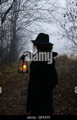 Die junge Frau geht durch den dunklen Wald, sie hält eine eiserne Laterne. Neblige Landschaft im Hintergrund. Stockfoto