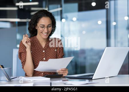 Junge schöne arabische Geschäftsfrau, die in einem modernen Büro mit Dokumenten, Papierkram arbeitet, Bericht liest und glücklich mit dem Ergebnis lächelt Stockfoto