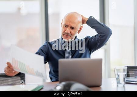 Ein besorgter Geschäftsmann sitzt hinter seinem Laptop und hält ein Papier in der Hand, während er im Büro arbeitet. Stockfoto