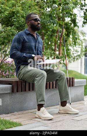 Vertikales Porträt eines blinden Mannes mit Sonnenbrille und Lesebuch in Blindenschrift im Freien Stockfoto