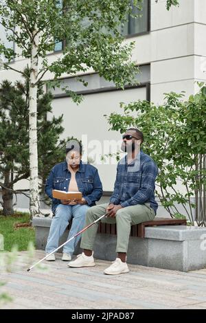 Vertikales Porträt eines erwachsenen Paares mit einem Partner mit Sehbehinderung, der zusammen auf einer Bank im Park sitzt Stockfoto