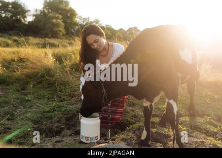 Die junge ukrainische Frau wässern das Kalb aus der Wasserkühl im traditionellen nationalen bestickten Hemd und Rock auf der Weide bei Sonnenuntergang. Ethnische ukrainische Nation Stockfoto