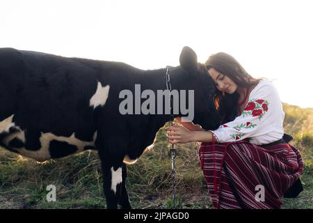 Die junge ukrainische Frau sitzt, lächelt und hält das Kalb in traditionellen nationalen bestickten Hemden und Rock auf der Weide. Ethnische ukrainische nationale cl Stockfoto