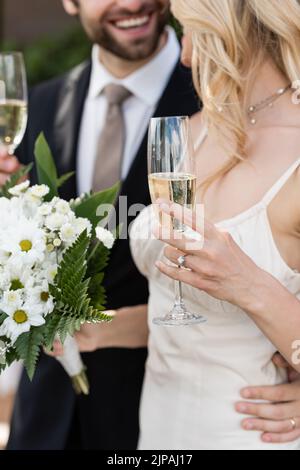 Verkürzte Ansicht der blonden Braut mit Blumenstrauß und Champagner in der Nähe des Bräutigams im Freien Stockfoto