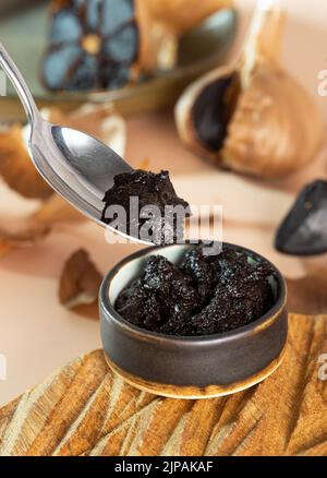 Eine kleine Tonschüssel mit schwarzer Knoblauchpaste auf einer handgefertigten Holzplatte. Löffel mit schwarzen Aioli. Fermentierte Knoblauchknolle im Hintergrund. Gesund Stockfoto