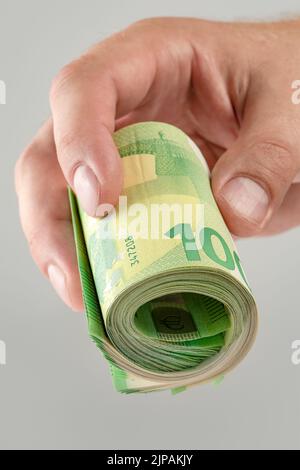 Die Hand hält eine Rolle von 100-Euro-Banknoten. Euro-Banknoten gerollt in der Hand eines weißen Mannes auf einem grauen Hintergrund. Das Konzept der finanziellen Unterstützung Stockfoto