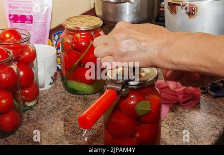 Der Prozess der Konservierung von Tomaten für den Winter. Frauenhände schließen die Deckel von Gläsern mit reifen roten saftigen Tomaten mit einem speziellen Schlüssel. Stockfoto