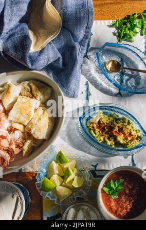 Sommer Meeresfrüchte Abendtisch verteilt, um Fisch Tacos, Garnelen, Guacamole, Limette, Tortilla, Salsa auf blauem und weißem Fischtischtuch in der Sonne Stockfoto