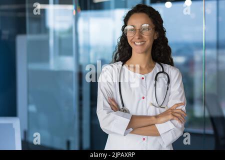 Porträt einer schönen lateinamerikanischen Ärztin, Ärztin bei der Arbeit in einem modernen Klinikbüro, stehend am Fenster in der Robe eines Bettlers, mit gekreuzten Armen lächelnd und mit Blick aus dem Fenster Stockfoto