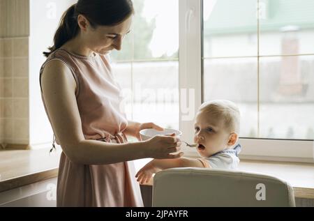 Mama füttert den Jungen Haferbrei mit einem Löffel von einem Teller. Essen Stockfoto