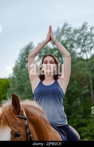Junge Frau macht Yoga auf einem Pferd vor dem Hintergrund von Bäumen Stockfoto