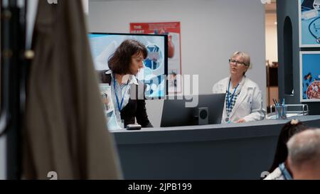 Krankenhausmitarbeiter, der am Empfang einen Termin vereinbart hat und dem Arzt und den Patienten im Wartezimmer administrative Informationen gibt. Medizinische Klinik Lobby mit Hilfe Zähler. Stockfoto