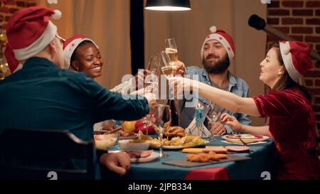 Festliche, vielfältige Menschen tragen traditionelle Hüte und knurren beim Weihnachtsessen mit Sektgläsern. Glückliche Familie, die den Winterurlaub mit hausgemachten Speisen und Sekt feiert. Handgeführte Aufnahme Stockfoto