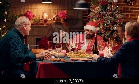 Lustige junge Erwachsene Person verkleidet als Weihnachtsmann diskutieren mit Menschen während der Feier Winterurlaub. Festlicher Mann trägt Weihnachtsmann Kostüm auf Weihnachtsessen Party im Gespräch mit Familienmitgliedern Stockfoto