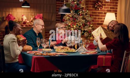 Fröhliche, multiethnische Familienmitglieder tauschen während des Weihnachtsessens Geschenke aus. Festliche, fröhliche Menschen schenken einander Geschenke, während sie den Winterurlaub gemeinsam zu Hause feiern. Stativaufnahme Stockfoto