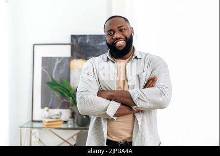 Positiv selbstbewusst schöner, erfolgreicher afroamerikanischer junger Mann in stilvoller Freizeitkleidung, mit kabellosen Kopfhörern, im Innenbereich mit gekreuzten Armen stehend, freundlich lächelnd auf die Kamera schauend Stockfoto