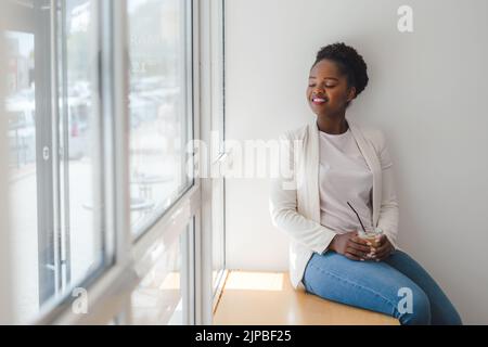 Lächelnde afroamerikanische Frau, die den Duft von Latte-Eiskaffee genießt und sich auf einem Fensterbrett eines Cafés entspannt. Eiskaffee Stockfoto