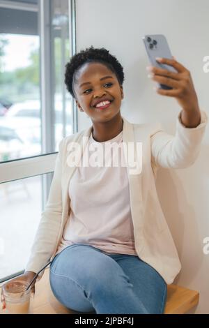 Porträt eines attraktiven afrikanischen Studenten, der eine Pause macht, um ein Selfie in einem Café zu machen. Frau, die einen Videoanruf auf dem Mobiltelefon macht, sitzt bei einem Stockfoto