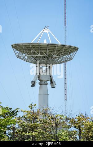 Parabolantennen-Satellitenschüssel vor dem Telekommunikationsturm mit einem klaren blauen Himmel im Hintergrund. Stockfoto