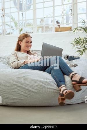 Entspannter, trendiger Designer, der in einer kreativen Startup-Agentur an einem Laptop arbeitet, während er auf einem Sitzsack sitzt. Coole, komfortable und glückliche Geschäftsfrau Stockfoto