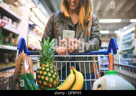 Ihre kleine Einkaufsassistentin. Eine Frau, die ein Mobiltelefon in einem Lebensmittelgeschäft benutzt. Stockfoto