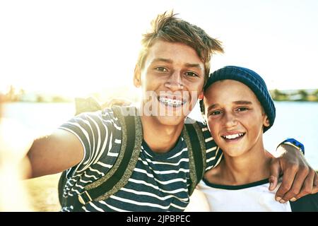 Sonnenschein sorgt immer für ein gutes Selfie. Beschnittenes Porträt zweier junger Brüder, die draußen Selfies mit einer Lagune im Hintergrund machen. Stockfoto