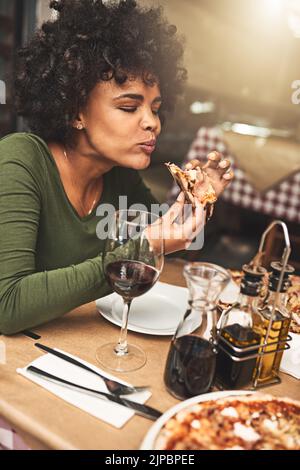 Diese Pizza ist die Bombe. Eine fröhliche junge Frau genießt ein Stück Pizza, während sie an einem Tisch im Restaurant sitzt. Stockfoto