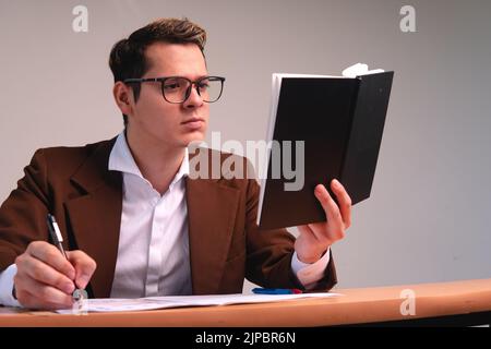 Geschäftsmann liest seinen schwarzen Notizblock. Kaukasischer und blonder Mann, der in einem braunen Anzug, einer Brille und an seinem Schreibtisch arbeitete. Mann, der in seinem Büro arbeitet. Hochwertige Fotos Stockfoto