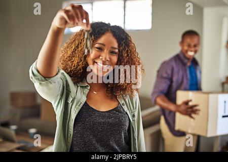 Die Schlüssel zu unserem Schloss. Porträt einer fröhlichen jungen Frau, die die Schlüssel zu ihrem Haus mit ihrem Partner in den hinteren Tragekisten hält. Stockfoto