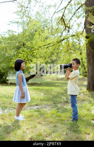 Netter kleiner asiatischer Junge, der sich wie ein professioneller Fotograf verhält, während er Fotos von seiner kleinen Schwester fotografiert Stockfoto