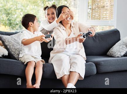 Familie, Videospiele und betrügerisches kleines Mädchen, das die Augen ihrer Mutter bedeckt, um ihren Bruder auf der Couch zu Hause gewinnen zu lassen. Unterhaltung, Joystick und mit Stockfoto