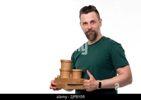 Der gutaussehende bärtige Umweltaktivist trägt ein grünes T-Shirt und hält umweltfreundliche Einwegkartons und Suppenbecher und zeigt den Daumen nach oben. Abfall Stockfoto