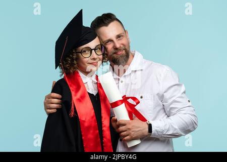 Smart whizzkid weibliche Kind in Abschlusskappe und Zeremonie Robe hält Zertifikat zeigt Zunge hat Spaß auf hellblauen Studio Hintergrund. Mein Vater gratuliert Stockfoto
