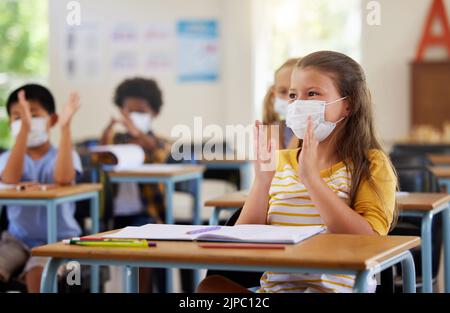 Sicherheit, Compliance und Bildung im Klassenzimmer in der Schule mit maskierten Schülern während der Corona-Pandemie. Junge Lernende klatschen, aufgeregt und Stockfoto