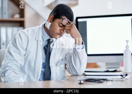 Gestresster, müder junger männlicher Arzt an seinem Schreibtisch in einem Krankenhaus. Arzt oder Gesundheitsmann erschöpft mit Schmerzen und Kopfschmerzen oder Wunde Augen am Stockfoto