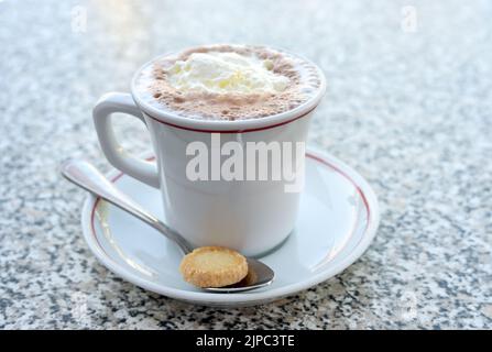Trinkschokolade mit Schlagsahne in einer weißen Tasse und einem Keks auf einem Löffel, süßes heißes Getränk auf einem Steintisch, Kopierraum, ausgewählter Fokus, enger Aufschlag Stockfoto