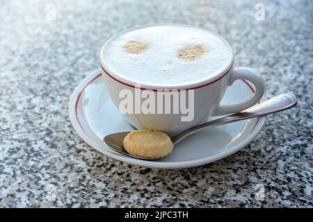 Cappuccino mit aufgeschäumter Milch in einer weißen Tasse und einem Keks auf einem Löffel, warmes Getränk auf einem Tisch aus Granit, Kopierraum, ausgewählter Fokus, schmale Tiefe Stockfoto