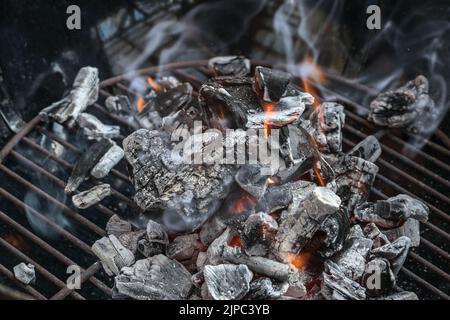 Glühende Holzkohle mit kleinen Flammen und Rauch in einem schwarzen Grill, Vorbereitung für einen Grill, Kopierraum, ausgewählter Fokus, enge Schärfentiefe Stockfoto