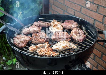 Steaks verschiedener Fleischsorten auf einem mobilen runden Grill werden auf einer Grillparty im Hinterhof, einem Kochkonzept im Freien, einem Kopierraum, über Holzkohle gebraten Stockfoto