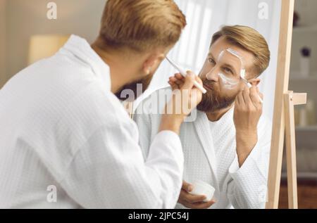 Hübscher erwachsener Mann in weißem Bademantel, der in einen Spiegel schaut und eine weiße Gesichtsmaske aus Lehm aufführt Stockfoto