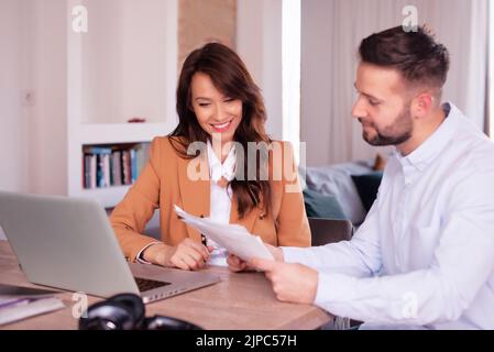 Geschäftsfrau und Geschäftsmann, die Dokumente betrachten, während sie hinter einem Laptop am Schreibtisch sitzen. Teamarbeit. Stockfoto