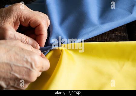 Weibliche alte ukrainische Frau Hände nähen die Nationalflagge der Ukraine gelb-blau am Tisch zu Hause in der Ukraine, Patriot, stoppen Sie den Krieg in der Ukraine 2022 Stockfoto