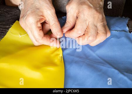 Weibliche alte ukrainische Frau Hände nähen die Nationalflagge der Ukraine gelb-blau am Tisch zu Hause in der Ukraine, Patriot, stoppen Sie den Krieg in der Ukraine 2022 Stockfoto