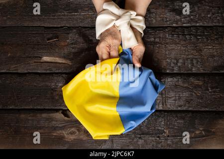 Hände einer ukrainischen Frau, mit einem weißen Seil gebunden, die eine gelbe und blaue Flagge der Ukraine auf einem hölzernen Hintergrund hält, Folter der Ukrainer an den Händen Stockfoto