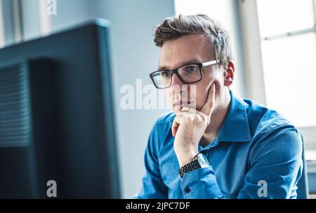 Hübscher junger Geschäftsmann mit Brille sitzt an seinem Tisch im Büro und schaut sich den Bericht auf seinem Computerbildschirm an. Stockfoto