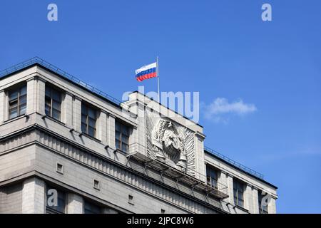 Parlamentsgebäude in Moskau mit russischer Flagge auf dem Hintergrund des blauen Himmels. Fassade der russischen Staatsduma mit sowjetischem Wappen, russische Autorität Stockfoto