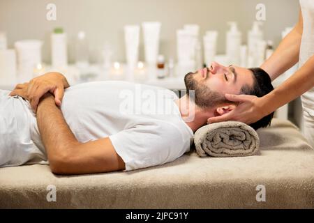 Seitenansicht eines entspannten jungen männlichen Klienten in einem weißen T-Shirt, das die Augen schließt und auf einem aufgerollten Handtuch liegt, während die Masseurin während der Spa-Zeit eine Nackenmassage macht Stockfoto