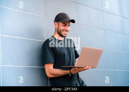 Der freundliche junge Mann in einem schwarzen T-Shirt und einer Mütze lächelt und stöbert in einem Netbook, während er sich an der gefliesten Wand eines modernen Gebäudes auf der Stadtstraße lehnt Stockfoto