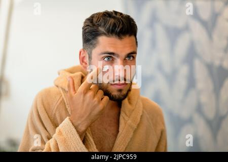 Junger bärtiger Mann im Bademantel, der während der morgendlichen Hautpflege auf den Spiegel schaut und feuchtigkeitsspendende Creme unter blauem Auge schmiert Stockfoto